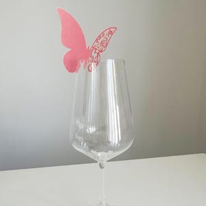 May include: A clear wine glass with a pink butterfly decoration. The butterfly has a detailed wing design and is positioned above the glass. The glass is on a white surface with a gray background.