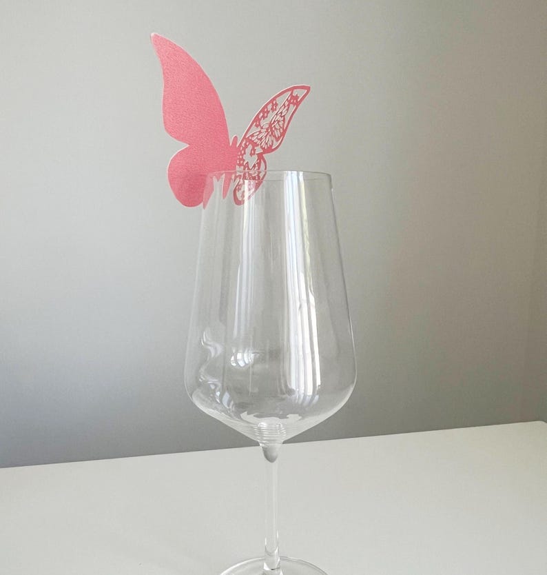 May include: A clear wine glass with a pink butterfly decoration. The butterfly has a detailed wing design and is positioned above the glass. The glass is on a white surface with a gray background.
