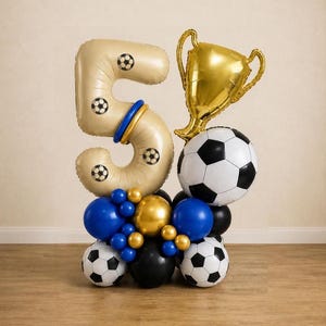 Puede incluir: Un arreglo de globos que presenta un globo grande de color beige con el número cinco decorado con patrones de balones de fútbol, un globo de trofeo dorado y un globo grande y redondo de fútbol. La base incluye globos de látex azules, dorados y negros.