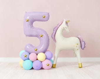 May include: A lilac number 5 balloon with gold star details, above a cluster of pastel balloons. A white unicorn balloon with a gold horn and hooves, and purple mane and tail, stands nearby. A party decoration.