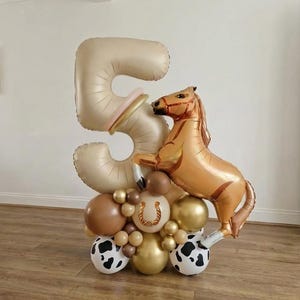 May include: A festive balloon arrangement for a fifth birthday. It features a large gold number 5, a rearing horse balloon, and a base of various balloons in gold, brown, and cow print. The arrangement is designed for a birthday celebration.