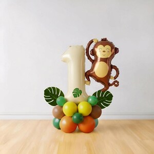 May include: A festive balloon arrangement for a first birthday celebration. It features a large number one balloon in cream, a brown monkey balloon, and a base of colorful balloons in green, yellow, brown, and orange, with green leaf accents.