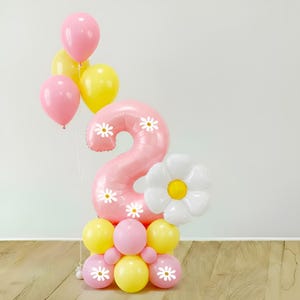 May include: A balloon arrangement featuring a large pink number two balloon decorated with white daisy prints. It is surrounded by smaller round balloons in shades of pink and yellow, plus a large white daisy-shaped balloon with a yellow center.