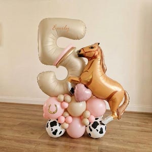 May include: A balloon arrangement for a fifth birthday. It features a large gold number 5 balloon with the name "Emily" in gold script. A gold horse balloon stands next to the number, with pink and cow-print balloons at the base.