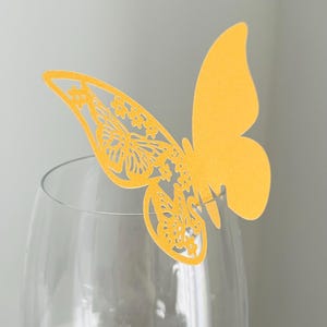 May include: A yellow butterfly-shaped decoration with intricate cut-out details, perched on the rim of a clear wine glass. The butterfly's wings feature floral patterns. The background is a soft, neutral color.