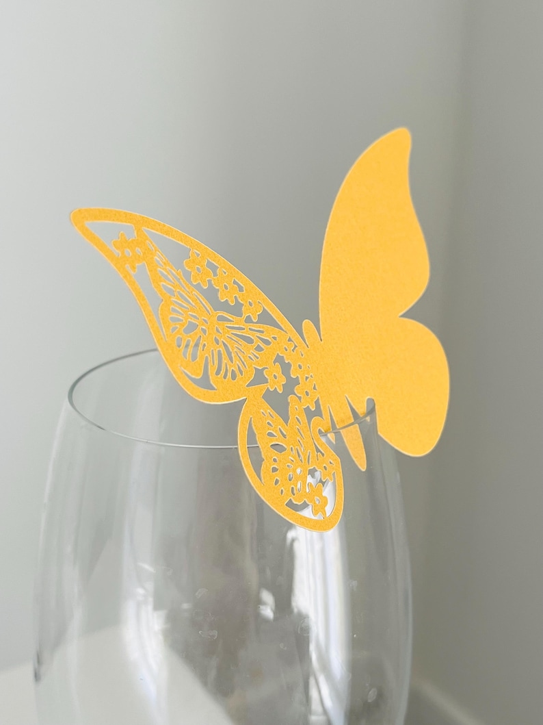 May include: A yellow butterfly-shaped decoration with intricate cut-out details, perched on the rim of a clear wine glass. The butterfly's wings feature floral patterns. The background is a soft, neutral color.