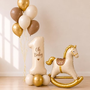 May include: A balloon arrangement featuring a cluster of gold, cream, and brown balloons, a large cream-colored number one balloon printed with the text Oh Baby and a heart, and a cream and gold rocking horse-shaped foil balloon.