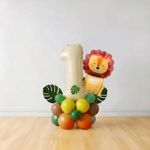 May include: A balloon arrangement for a first birthday celebration. It features a large beige number one balloon, a lion-shaped balloon, and a base of colorful balloons in shades of green, brown, and orange. Decorative green leaves are also included.
