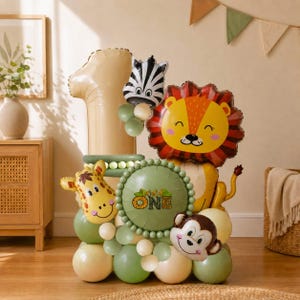 May include: A balloon arrangement featuring a large beige number one, a round green balloon with the text Wild One, and foil balloons shaped like a lion, zebra, giraffe, and monkey. The base is constructed from various shades of green and cream balloons.