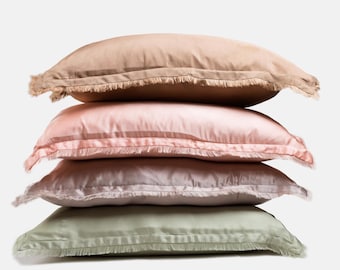 May include: A stack of four decorative pillows in shades of beige, pink, and green. Each pillow has a rectangular shape with a fringed edge. The pillows are stacked on top of each other against a white background.