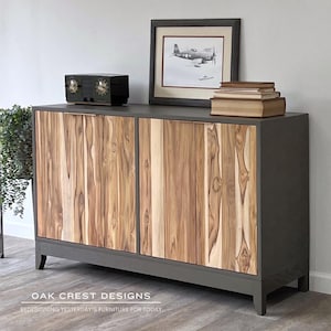 May include: A gray cabinet with two doors featuring vertical wood paneling. The cabinet has a dark gray frame and legs. On top are a vintage radio, framed airplane art, and books. The text "OAK CREST DESIGNS" and "REDESIGNING YESTERDAY'S FURNITURE FOR TODAY" are at the bottom.