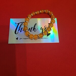 May include: An orange beaded bracelet with silver accents rests on a holographic business card. The card reads "Thank you" in cursive, with the text "for supporting my small business" below. The background is a solid red.