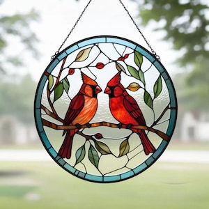 May include: A round stained glass window featuring two red cardinal birds perched on a branch with green leaves and red berries. The window has a blue border and is suspended by a chain.