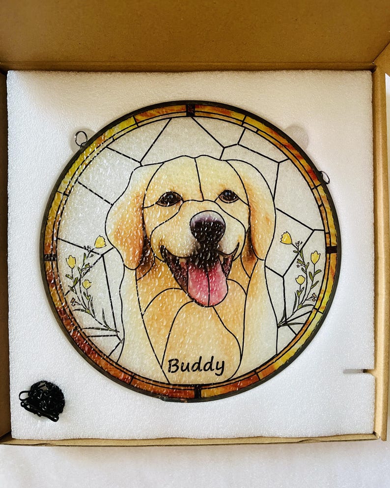 May include: Round stained glass window featuring a smiling golden retriever dog with the name "Buddy" at the bottom. The dog is in shades of tan and white, with a pink tongue. The window has a yellow and brown border.