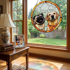 May include: A stained glass window featuring two dogs, a pug and a Labrador, with the names "Gordo" and "Baker" above them. The window is round with a gold border and hangs in front of a window overlooking a green lawn.