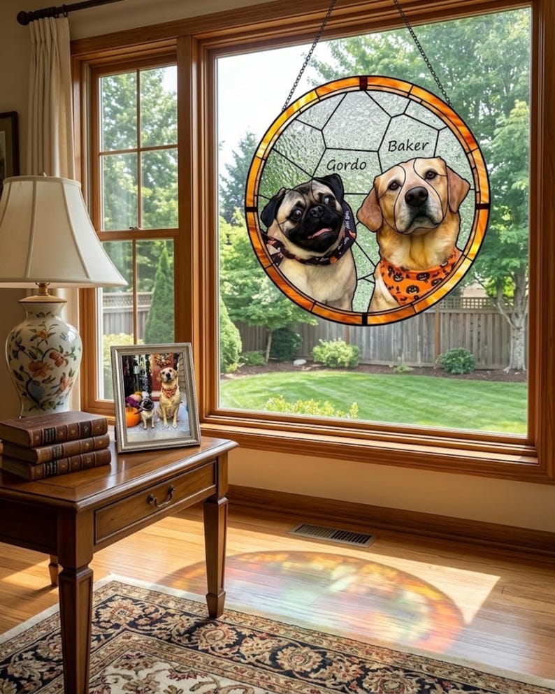 May include: A stained glass window featuring two dogs, a pug and a Labrador, with the names "Gordo" and "Baker" above them. The window is round with a gold border and hangs in front of a window overlooking a green lawn.
