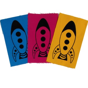 Peut inclure: Trois patchs en tissu avec un motif de fusée noire. Les patchs sont bleus, roses et jaunes.
