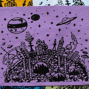 Puede incluir: Un cuadrado de tela morado con una ilustración en blanco y negro de una ciudad espacial con planetas, estrellas y cohetes. La ciudad está situada en una superficie rocosa.