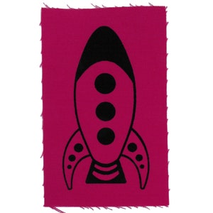 May include: A black rocket ship silhouette on a pink fabric patch. The rocket has three round windows and a striped tail.