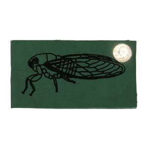 May include: A green fabric patch with a black silhouette of a cicada insect. The patch has a silver button on the top right corner.