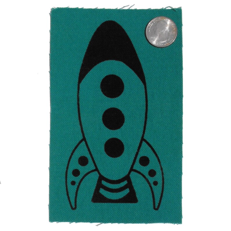 May include: A teal fabric patch featuring a black rocket ship design. The rocket ship has three black circles in the centre and a black stripe at the bottom.