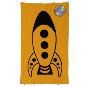 May include: A yellow fabric patch featuring a black rocket ship design. The rocket ship has three black circles in the centre and a black stripe at the bottom.