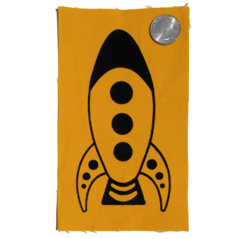 May include: A yellow fabric patch featuring a black rocket ship design. The rocket ship has three black circles in the centre and a black stripe at the bottom.