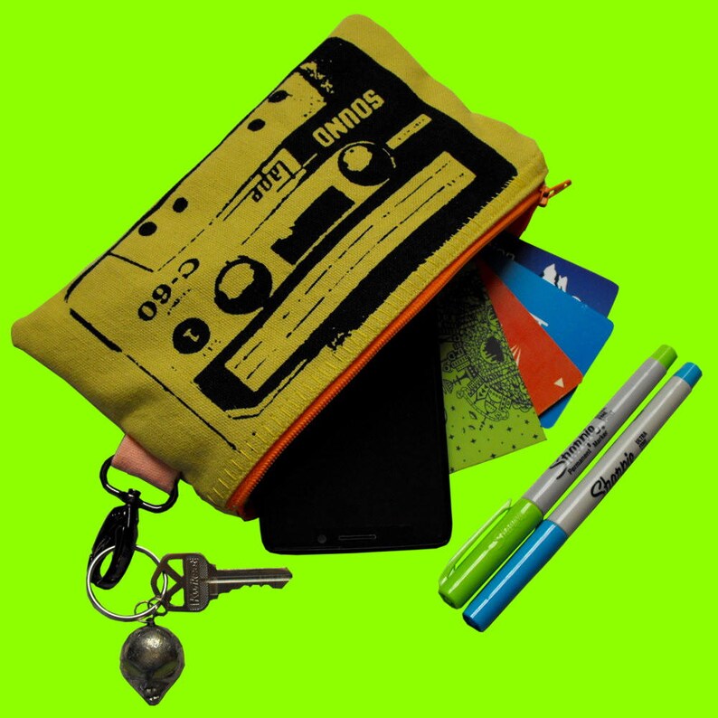 May include: A yellow canvas zippered pouch with a black and white cassette tape design. The pouch is open and has a black phone, a green and black marker, and a silver marker inside. The pouch has a black zipper pull with a silver key ring attached.