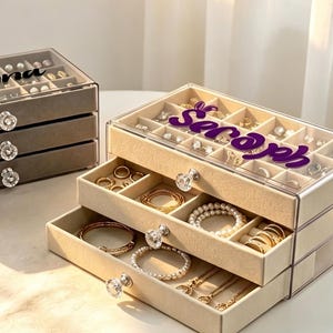 May include: Two rectangular jewellery organisers with clear acrylic frames and velvet-lined drawers. The left organiser is grey with the name Brynna in black script. The right organiser is beige with the name Seraph in purple script.