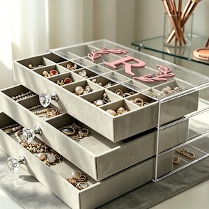 May include: A clear acrylic jewelry organizer with three pull-out drawers lined in soft gray velvet. The top drawer features a personalized pink letter R surrounded by a decorative leaf design. Each drawer has a round, faceted crystal-style knob.