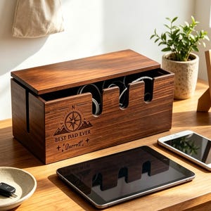 Peut inclure: Une boîte de rangement pour câbles en bois foncé rectangulaire avec un couvercle amovible, présentant une gravure laser d'une boussole, le texte "BEST DAD EVER" et "Darrell" sur le devant, et plusieurs découpes pour les câbles.