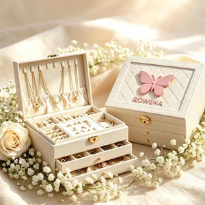 May include: A cream-colored jewelry box with a quilted lid featuring a pink butterfly and the name ROWENA. The open box reveals multiple compartments, gold-toned metal latches, and various gold chains, rings, and earrings stored inside.