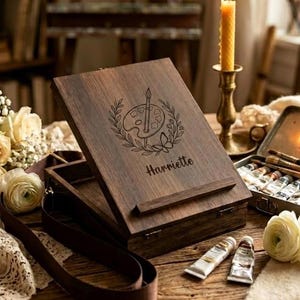 Puede incluir: Una caja de artista de madera oscura con tapa con bisagras, grabada con un diseño de paleta y pincel y el nombre "Harriette". La caja está abierta, revelando un compartimento interior. Se ven tubos de pintura y pinceles.