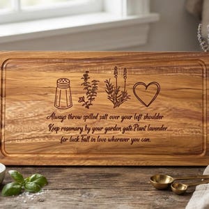 May include: A rectangular wooden cutting board with a juice groove and laser-engraved illustrations of a salt shaker, rosemary, lavender, and a heart. The text reads: Always throw spilled salt over your left shoulder. Keep rosemary by your garden gate. Plant lavender for luck. Fall in love wherever you can.
