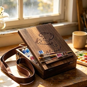 Op de afbeelding: Een houten kunstenaarsdoos met een bruine leren riem, open om verftubes, aquarelverf en penselen te onthullen. Het deksel van de doos is gegraveerd met een palet, een verftube en de naam "Manuel". Een mok en een aquarelpalet staan in de buurt.