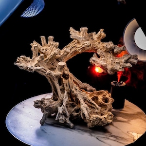 May include: A beige, driftwood-style decorative sculpture with a tree-like form. The sculpture has a rough, textured surface and is placed on a marble-patterned surface. A small, black cylindrical object with a red light is next to the sculpture.