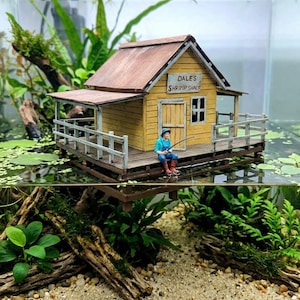 May include: A miniature yellow "Dale's Shrimp Shack" with a brown roof and porch, set in an aquarium. A small figure is fishing from the porch. The aquarium features aquatic plants, driftwood, and a sandy substrate.