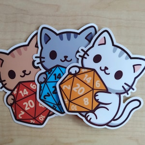 Puede incluir: Tres pegatinas de gatos, cada una sosteniendo un dado de veinte caras. Los gatos son en tonos naranja, gris y blanco. Los dados son rojos, azules y naranjas, con números blancos. Un artículo divertido para los entusiastas de los juegos de mesa.