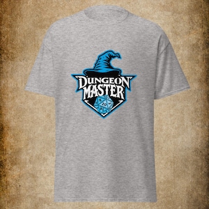 Puede incluir: Una camiseta gris con un sombrero de mago azul y las palabras "DUNGEON MASTER" en una fuente estilizada. Debajo del texto hay un dado azul de veinte caras. El diseño está centrado en la parte delantera de la camiseta.