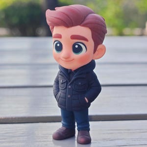 Op de afbeelding: Een cartoonfiguur van een jonge persoon met rood haar, gekleed in een marineblauwe jas, een blauwe spijkerbroek en bruine schoenen. De figuur heeft grote blauwe ogen en glimlacht. Het beeldje staat op een houten oppervlak.