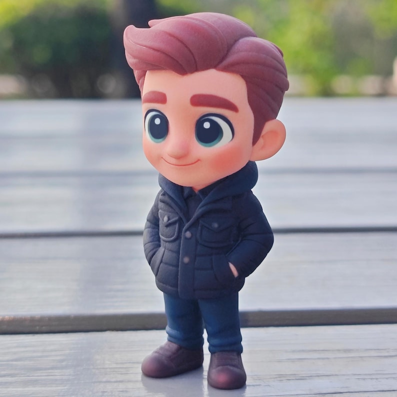 Op de afbeelding: Een cartoonfiguur van een jonge persoon met rood haar, gekleed in een marineblauwe jas, een blauwe spijkerbroek en bruine schoenen. De figuur heeft grote blauwe ogen en glimlacht. Het beeldje staat op een houten oppervlak.