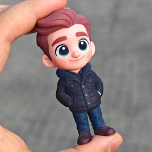 Op de afbeelding: Een beeldje van een stripfiguurjongen met rossig haar, gekleed in een marineblauwe jas, een blauwe hoodie, een blauwe spijkerbroek en bordeauxrode schoenen. De jongen heeft grote, expressieve ogen en een lichte glimlach. Het beeldje wordt in een hand gehouden.