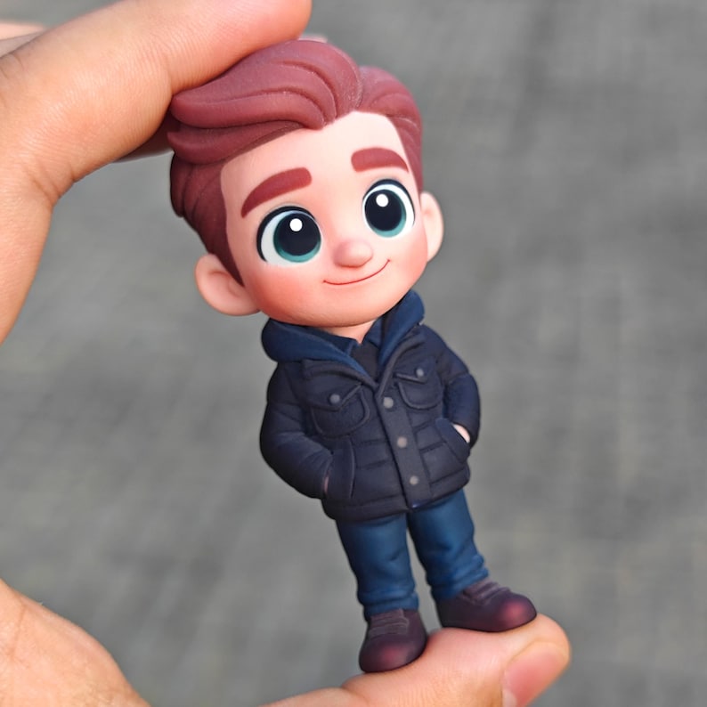 Op de afbeelding: Een beeldje van een stripfiguurjongen met rossig haar, gekleed in een marineblauwe jas, een blauwe hoodie, een blauwe spijkerbroek en bordeauxrode schoenen. De jongen heeft grote, expressieve ogen en een lichte glimlach. Het beeldje wordt in een hand gehouden.