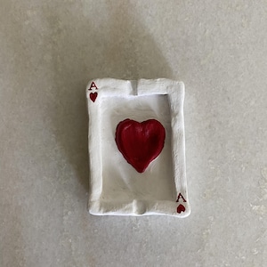 Puede incluir: Cenicero de cerámica blanca con forma de carta de juego as de corazones. El corazón está pintado de rojo.