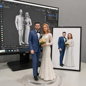 May include: A 3D printed figurine of a couple in wedding attire, featuring a person in a blue suit and a person in a white lace gown holding a bouquet. The figurine stands on a clear circular base, with a computer monitor showing a 3D model and a framed photo.