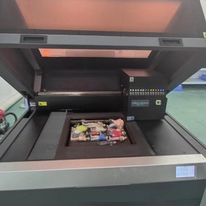 May include: A large industrial UV flatbed printer with its top lid open, revealing a black printing bed containing several small plastic human figurines. The machine features a metallic silver and black exterior with a control panel and ink system.