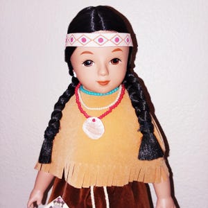 May include: A Native American doll with long black braids, wearing a brown suede dress with fringe, a white headband with pink and red geometric designs, and a necklace with turquoise and red beads.