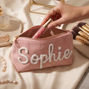 Puede incluir: Un neceser de maquillaje de pana rosa empolvado con el nombre Sophie bordado en letras blancas cursivas en la parte delantera. El estuche tiene una cremallera dorada y se muestra siendo llenado con un tubo de brillo labial rosa.