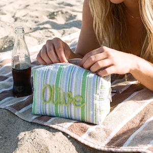 May include: A rectangular zippered pouch featuring a vertical striped pattern in shades of green, white, and purple. The word Olive is embroidered in a cursive green font on the front of the pouch. A glass bottle containing a dark liquid sits nearby.