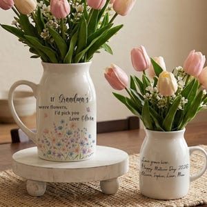 May include: Two white ceramic vases filled with pink and cream-colored tulips and greenery. One vase is on a small, round, white wooden stand. The vases have black text that reads "If Grandma's were flowers, I'd pick you Love Olivia" and "Love grows here: Happy Mother's Day 2026 Olivia, Sophie, Liam, Mia."